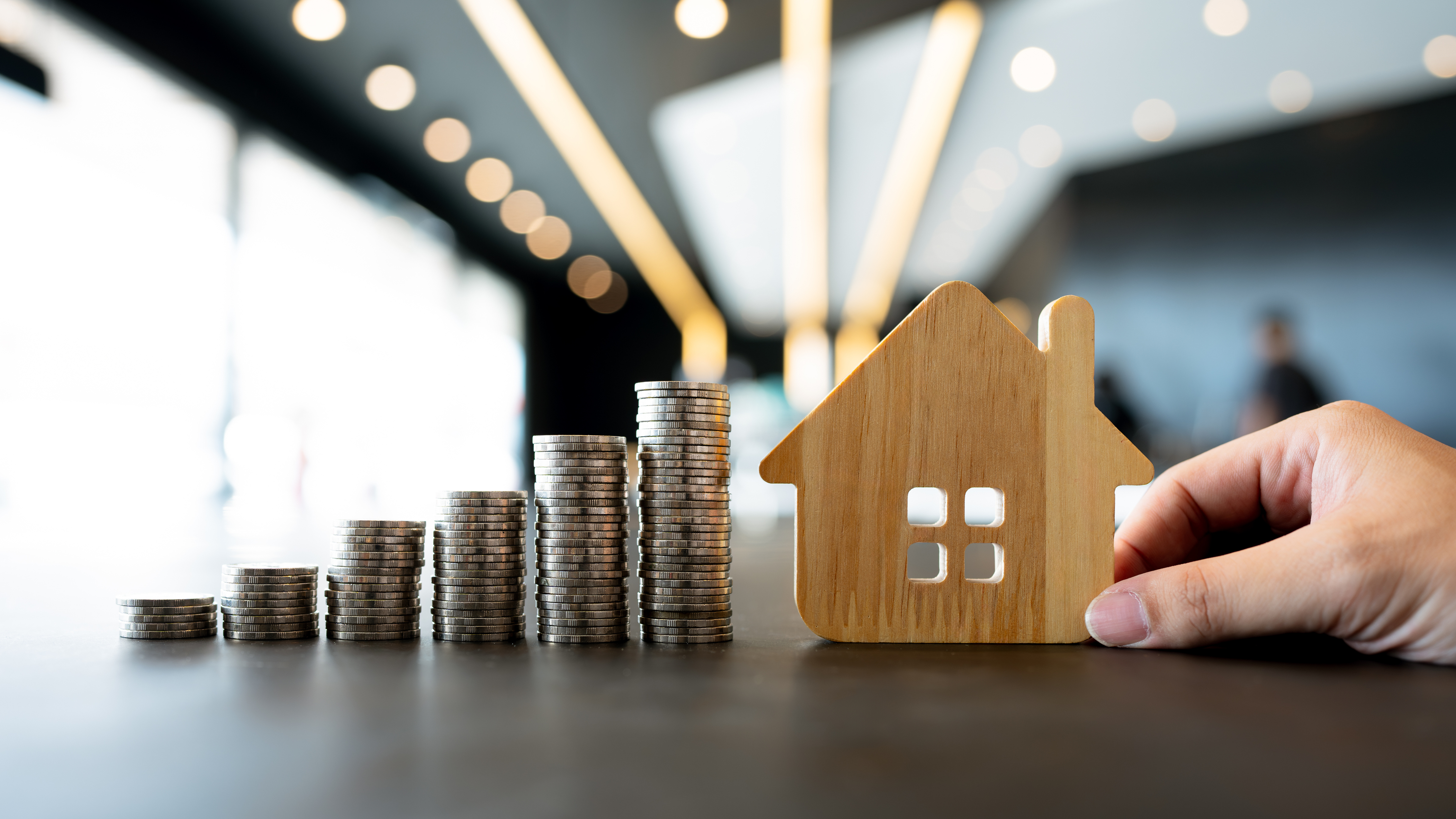 stacked-coins-and-hand-holding-wooden-house-icon-on-table